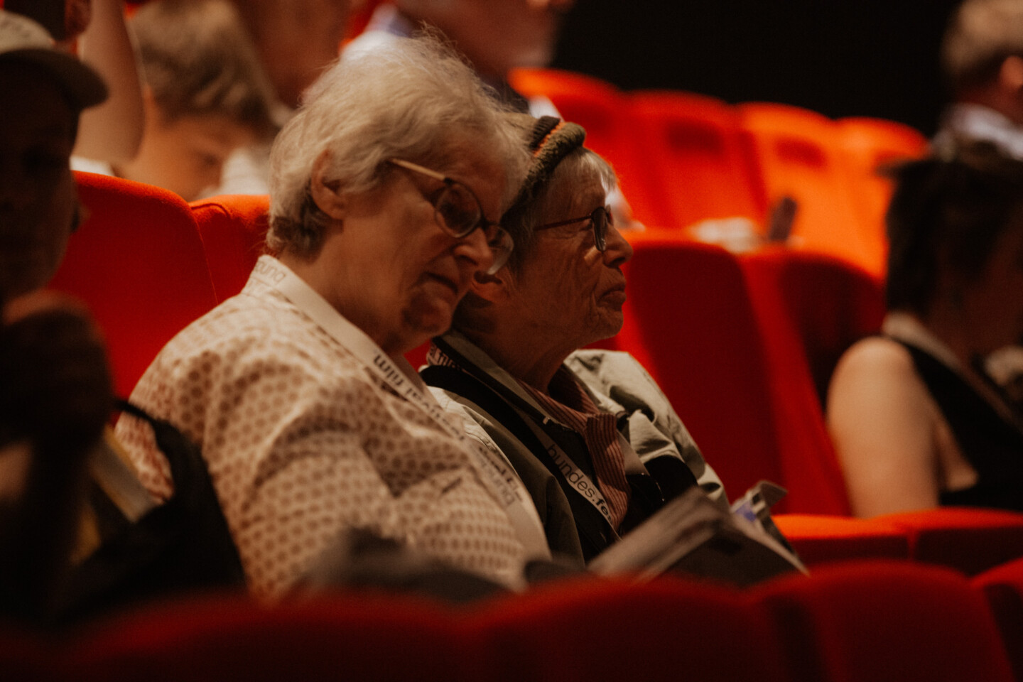 Zwei ältere Frauen sitzen auf roten Kinosesseln. Die linke von beiden blättert in einer Broschüre