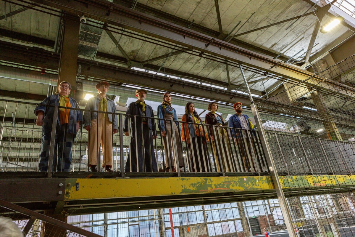 Sieben Personen verschiedenen Alters in Kleidung im Stil der 1970er Jahre stehen auf einer Empore in einem alten Fabrikgebäude.