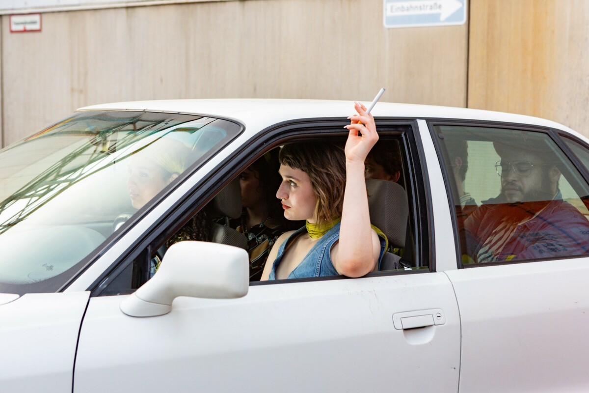 Eine junge Frau sitzt auf dem Fahrersitz eines weißen Autos älteren Baujahrs und hält eine Zigarette aus dem geöffneten Fenster.
