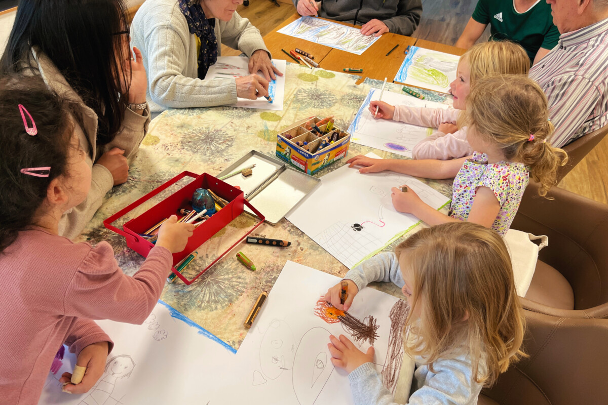 Eine Gruppe von alten Menschrn und Kindern, die um einen Tisch sitzen und zeichnen