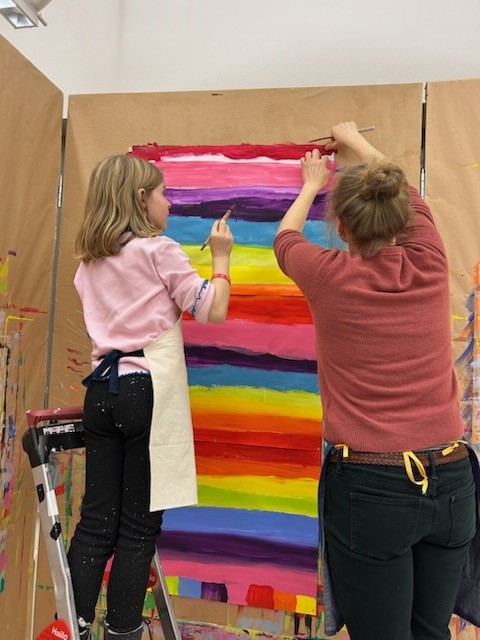 Eine Frau und ein Mädchen malen bunte Querstreifen an eine Wand mit Packpapier.