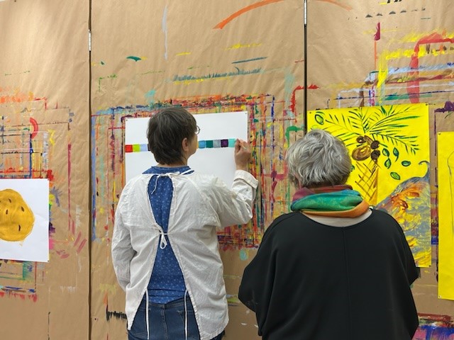 Zwei ältere Frauen stehen vor einer mit Packpapier bespannten Wand, an der weiße und gelbe Papierbögen mit Gemälden darauf hängen. Eine von ihnen malt an einem der Gemälde weiter.