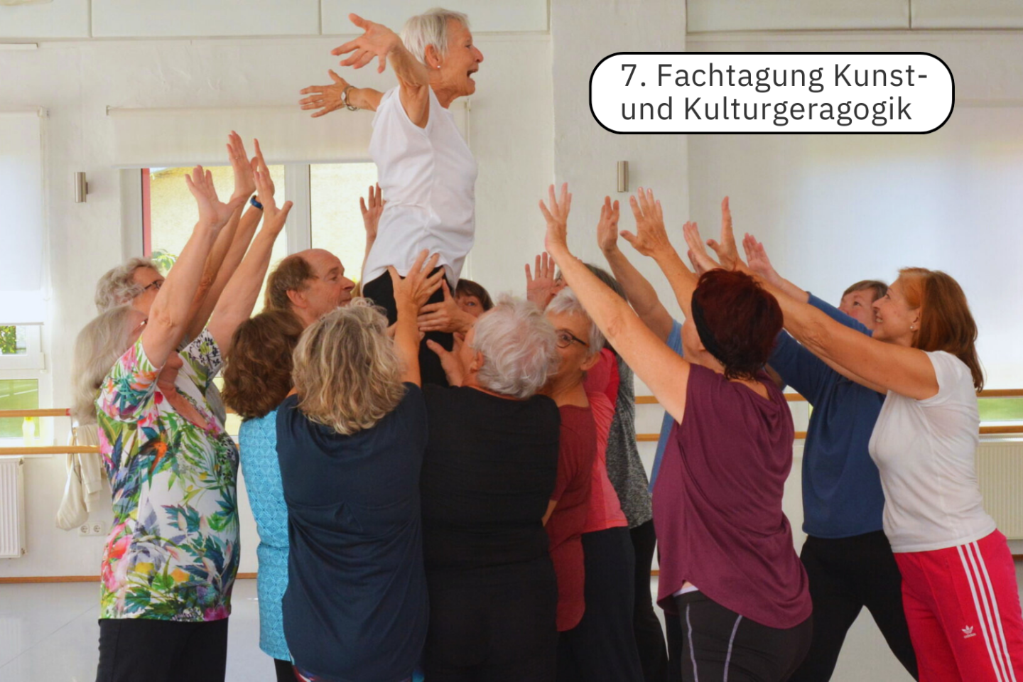 Foto einer Gruppe von Menschen, die eine ältere Frau in die Luft heben, mit dem Schriftzug „7. Fachtagung Kunst- und Kulturgeragogik“ darauf.