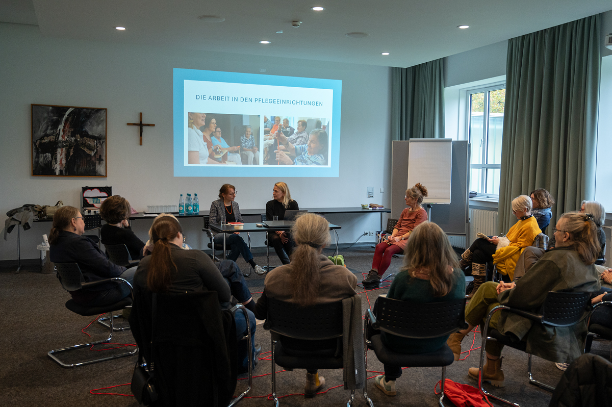 Eine Gruppe von Personen sitzen auf Stühlen im Kreis. An der Wand über ihnen sind auf einer Projektionsfläche Fotos von älteren und Jüngeren Menschen zu sehen sowie der Text „Die Arbeit in den Pflegeeinrichtungen“.