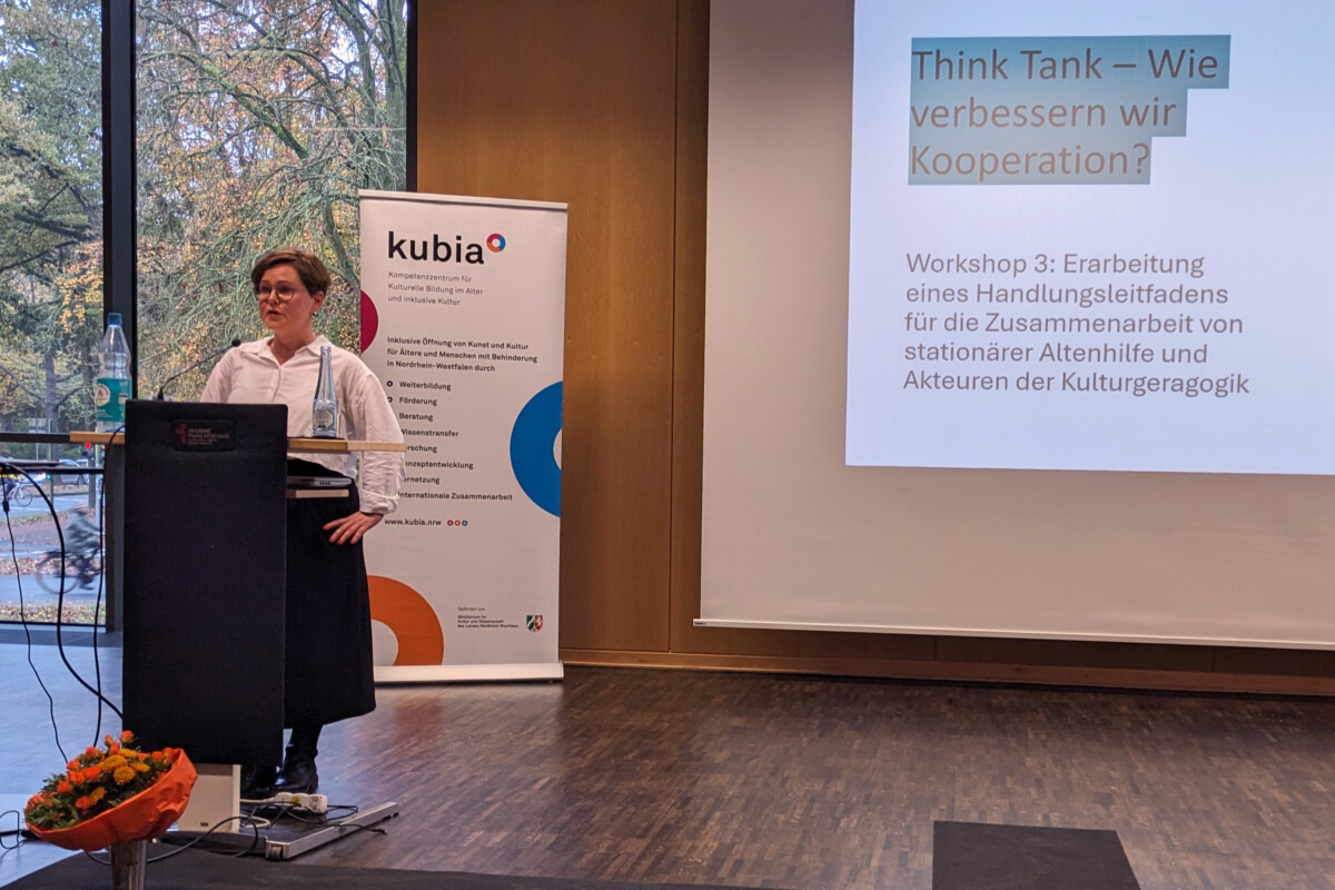 Nina Lauterbach-Dannenberg steht hinter einem Rednerpult. Hinter ihr steht auf einer Projektionsfläche: „Think Tank – Wie verbessern wir Kooperation? Workshop 3: Erarbeitung eines Handlungsleitfadens für die Zusammenarbeit von stationärer Altenhilfe und Akteuren der Kulturgeragogik“.