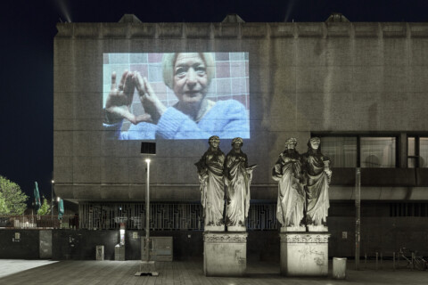 Eine nächtliche Szene mit einer großen Außenprojektion einer Frau, die eine rautenförmige Handbewegung macht, auf der Fassade eines modernen Betongebäudes. Im Vordergrund stehen vier klassische Statuen auf Sockeln.