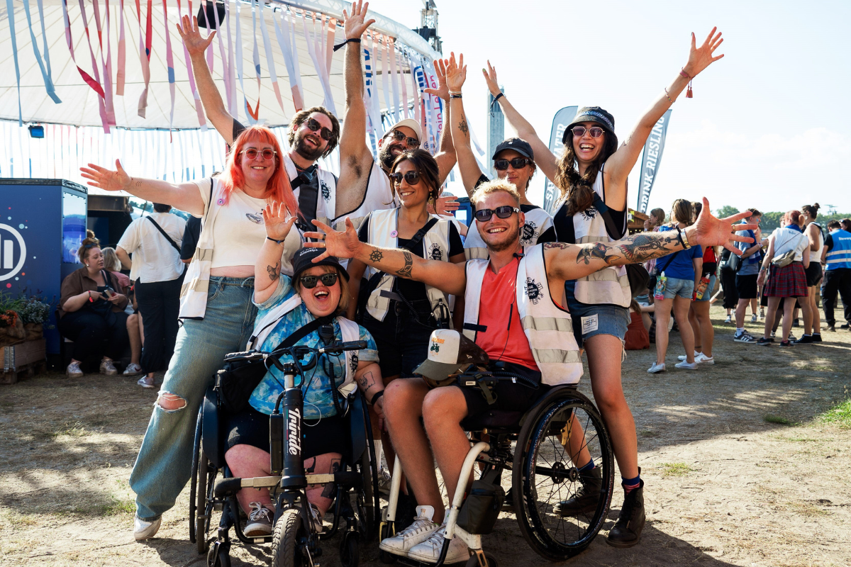 Eine Gruppe von acht Personen posiert mit hochgereckten Armen im Freien auf einem Festival, alle tragen weiße Westen mit Logos von „WIR kümmern uns“. Zwei Personen sitzen in Rollstühlen, während die anderen andere um sie herum stehen oder hocken. Im Hintergrund steht eine Art großes Zelt mit bunten Flatterbändern daran.