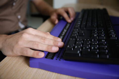 Eine Person nutzt eine Braillezeile vor einer herkömmlichen schwarzen Tastatur. Die linke Hand tastet die erhabenen Braille-Zeichen, während die rechte Hand auf der Tastatur ruht. Die Braillezeile ist lila und liegt auf einem Holztisch.