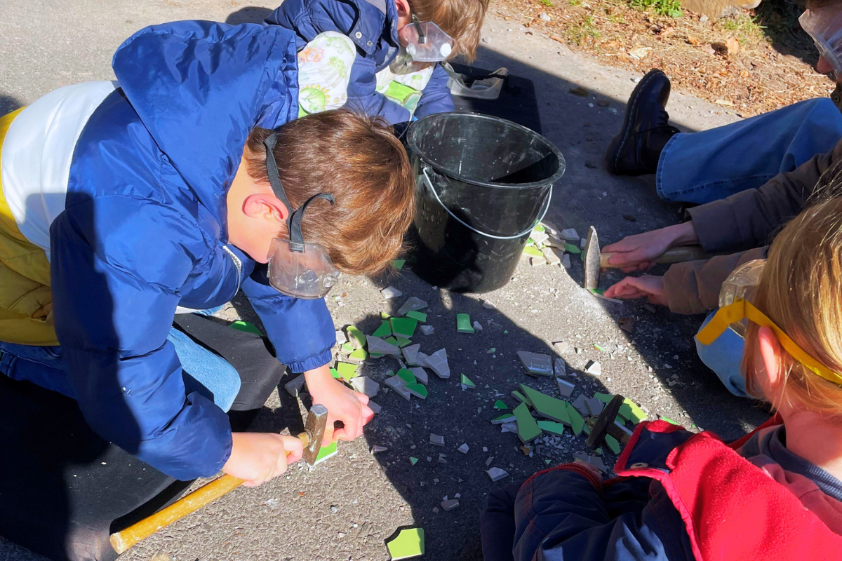 Mehrere Kinder, die Schutzbrillen tragen, zerschlagen mit Hämmern bunte Fliesen auf dem Boden.