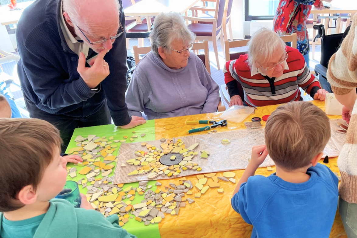 Kinder und Senior*innen sitzen gemeinsam an einem Tisch und gestalten ein Mosaik. Ein älterer Mann mit Brille lehnt sich nachdenklich über das Werk, während eine Seniorin und ein weiterer älterer Mann im Sitzen mitarbeiten. Die Kinder sitzen ihnen gegenüber.