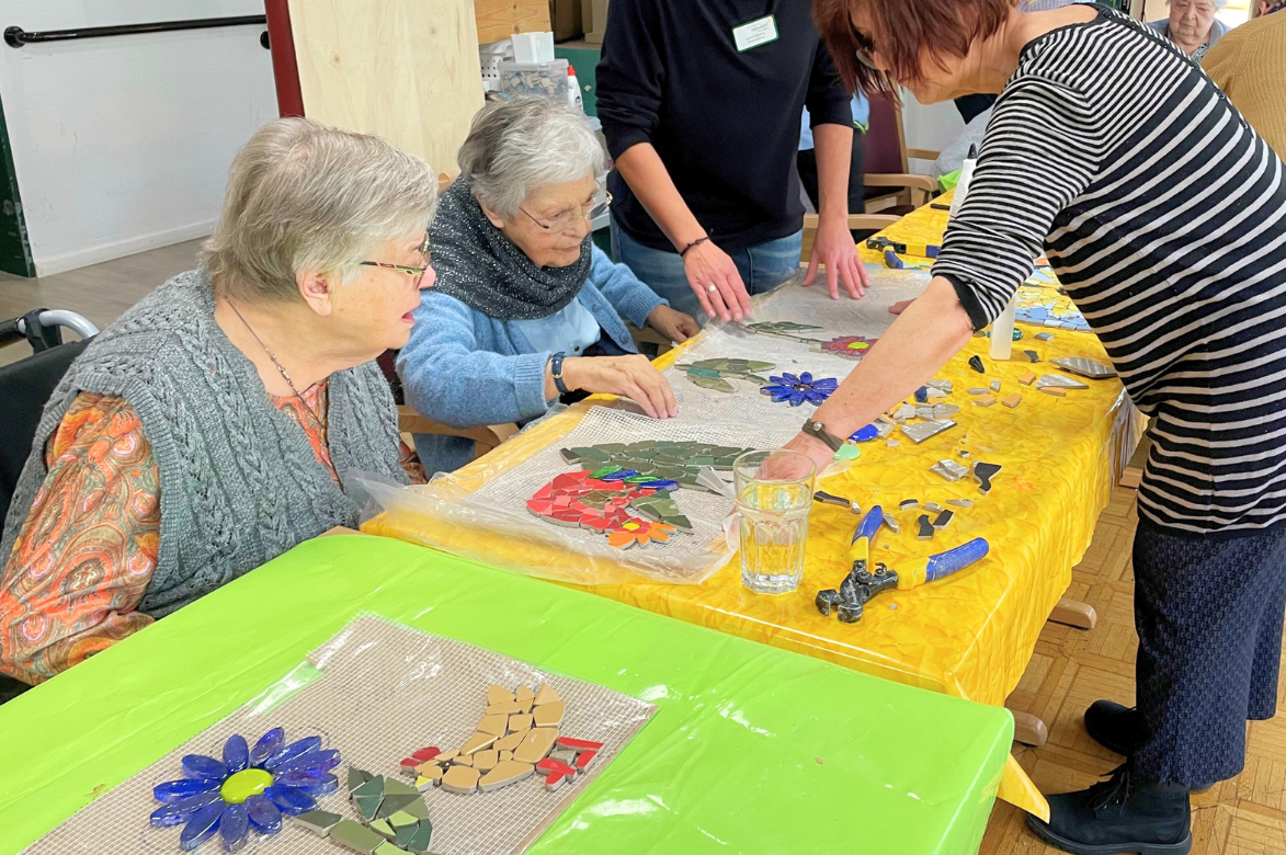 Zwei ältere Frauen, sitzen an einem Tisch, auf dem Mosaikmotive liegen. Neben ihnen stehen zwei weitere Frauen und arbeiten an den Mosaiken.