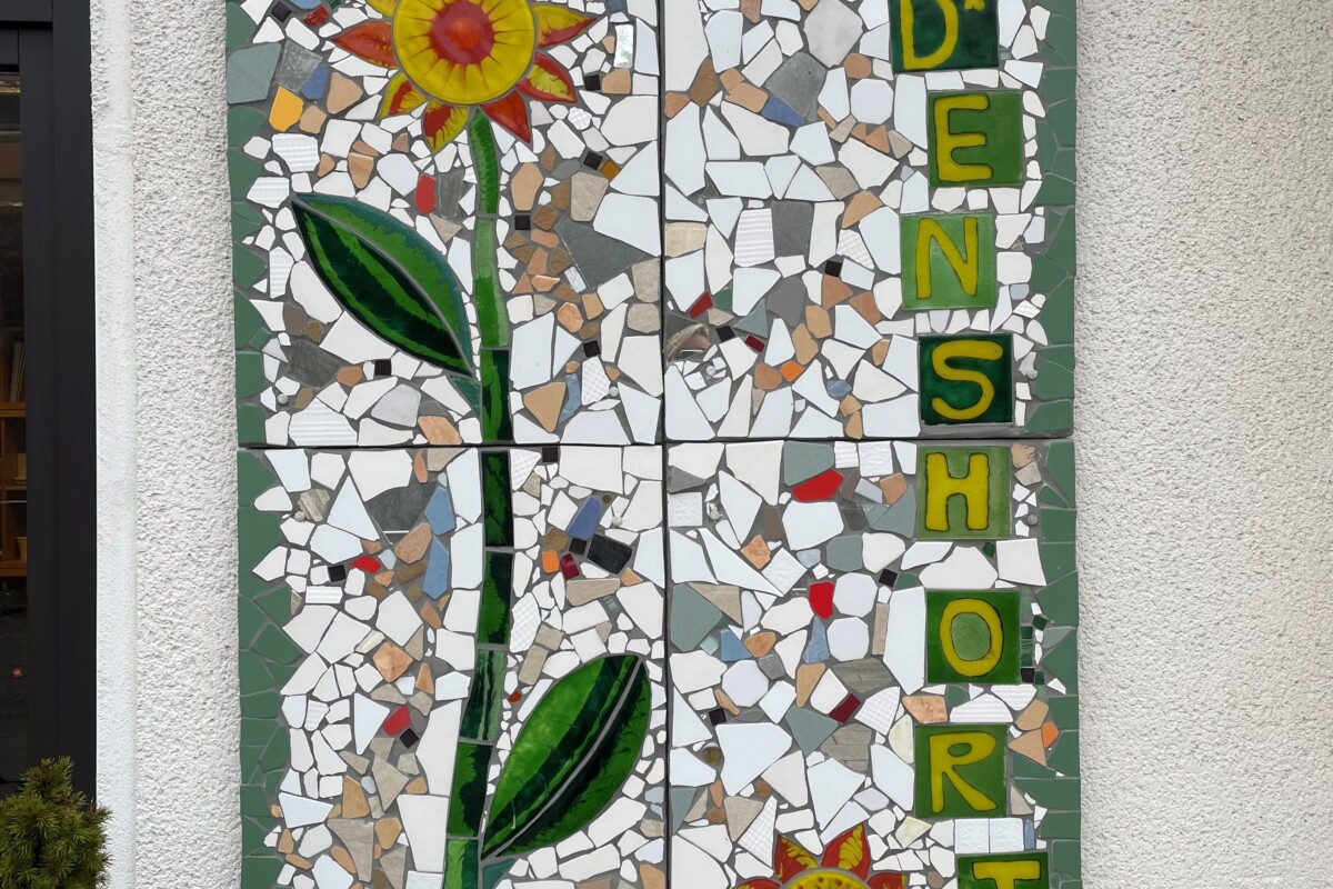 Das Bild zeigt ein großes Mosaik an einer Außenwand, das einen stilisierten Sonnenblumenstängel mit Blättern und gelb-roten Blüten darstellt. Das Mosaik besteht aus unregelmäßig geformten Fliesenstücken in Weiß, Beige und Grautönen, eingerahmt von einem grünen Rand. Rechts neben der Sonnenblume steht vertikal das Wort „FRIEDENSHORT“ in grünen Buchstaben auf gelbem Hintergrund.