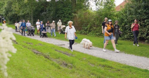 Menschen unterschiedlichen Alters und mit Kopfhören gehen auf einem Weg neben einer Wiese. Eine Person führt einen weißen Hund an der Leine links neben sich.