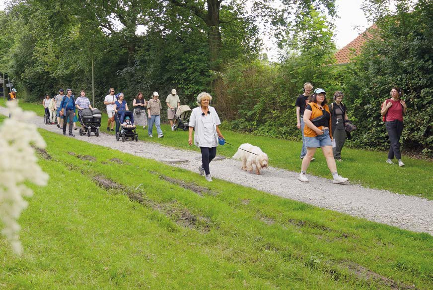 Menschen unterschiedlichen Alters und mit Kopfhören gehen auf einem Weg neben einer Wiese. Eine Person führt einen weißen Hund an der Leine links neben sich.